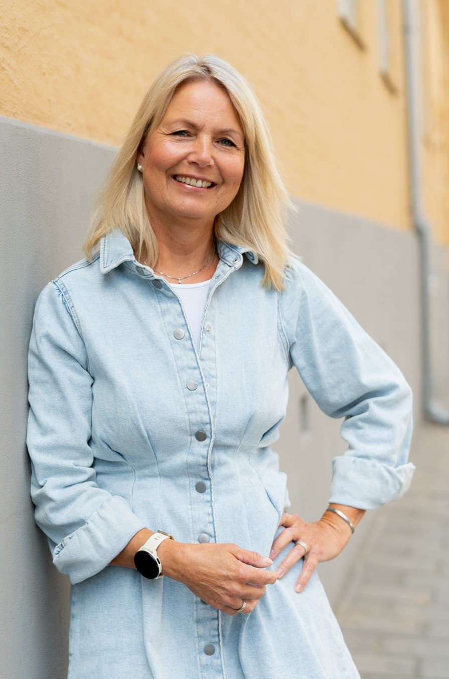Kvinne står inntil en vegg og holder hånda på hofta mens hun ser inn i kameraet og smiler.